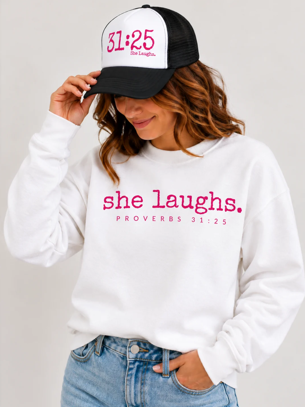 Woman wearing a white She Laughs sweatshirt with hot pink lettering and a 31:25 trucker hat