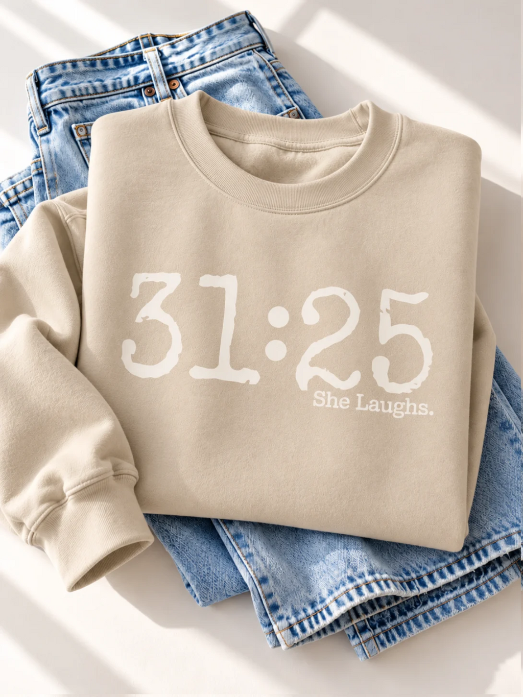 Sand 31:25 sweatshirt with large ivory front graphic, folded beside light-wash denim jeans on a bright background.