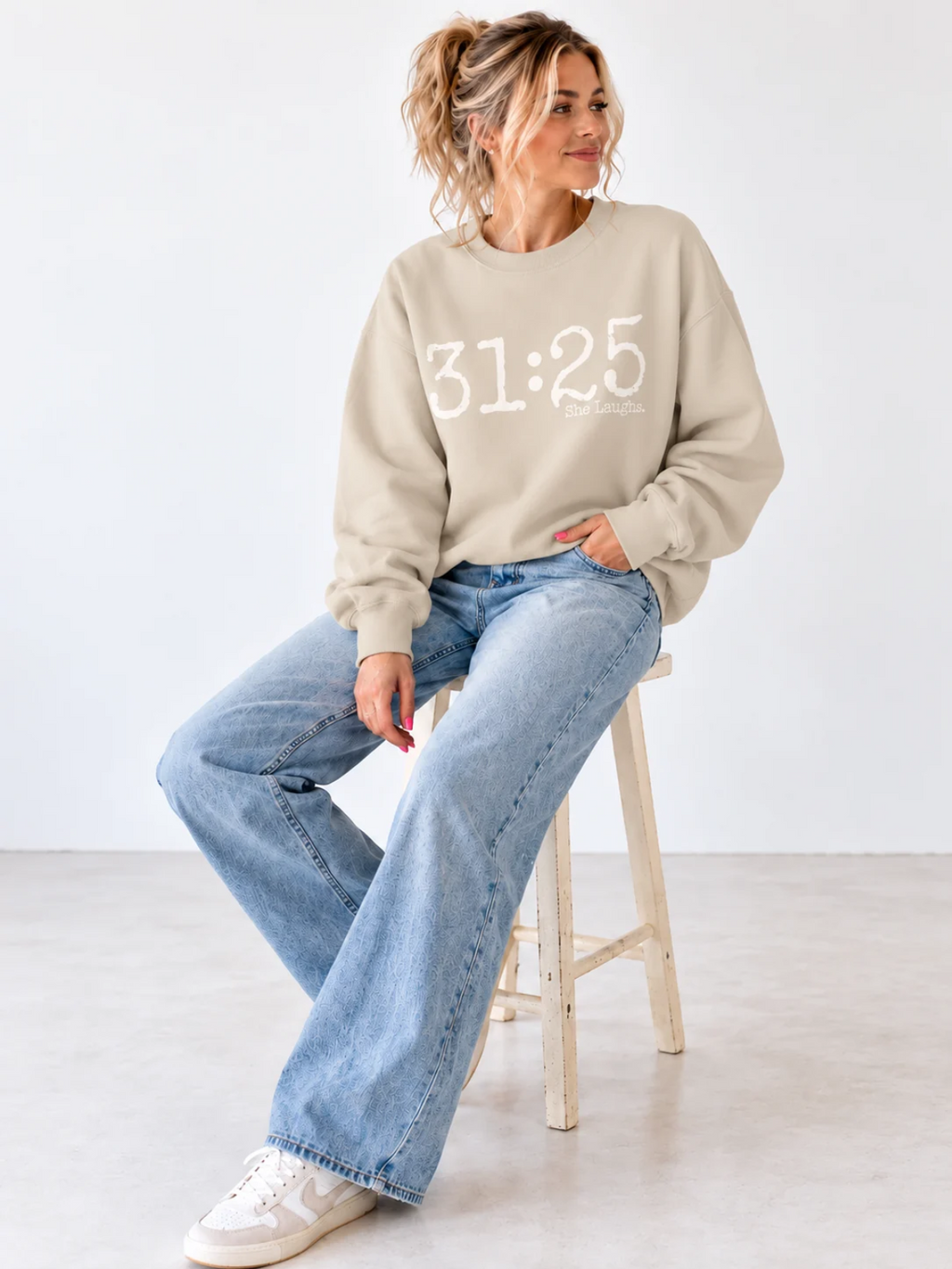 Blonde woman seated on a stool wearing a Sand 31:25 crewneck sweatshirt with light-wash jeans and sneakers.