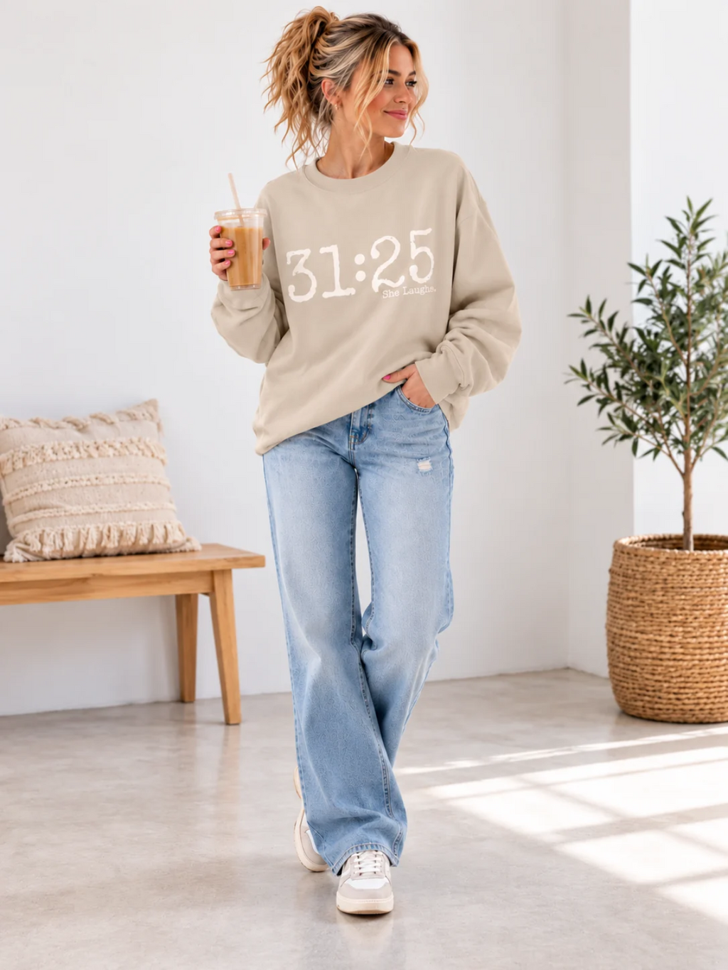 Woman standing in a Sand 31:25 sweatshirt, relaxed blue jeans and white sneakers in a bright studio.