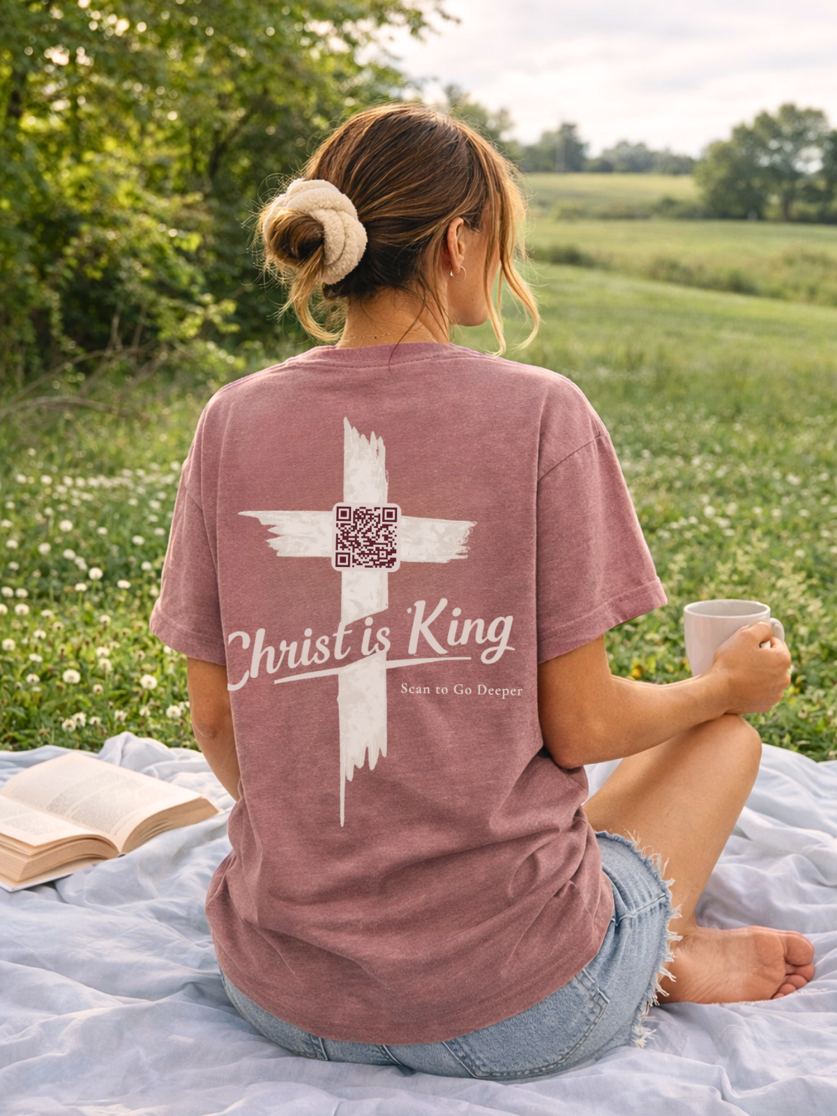 Christian Woman wearing a 'Christ is King' t-shirt with a cross design, sitting in a field holding a cup.