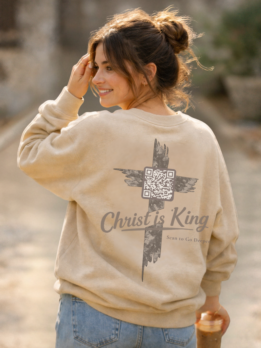 Woman wearing a beige sweatshirt with 'Christ is King' text and cross design. John 12-12:15