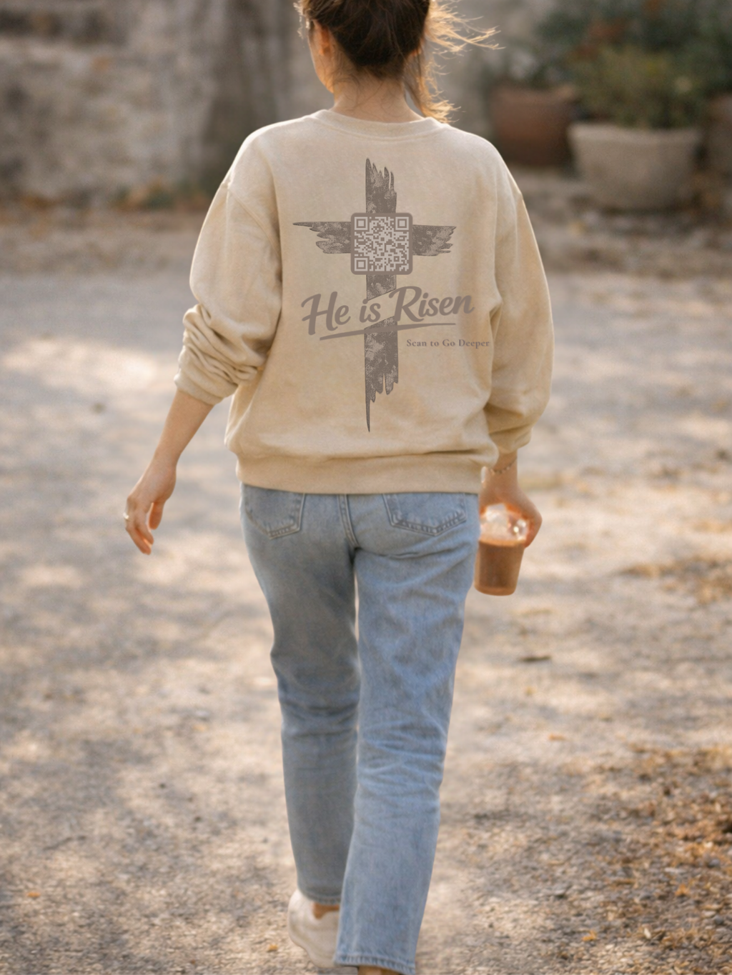 Christian Woman wearing a beige sweatshirt with 'He is Risen' text and cross design, walking outdoors. Matthew 28:6