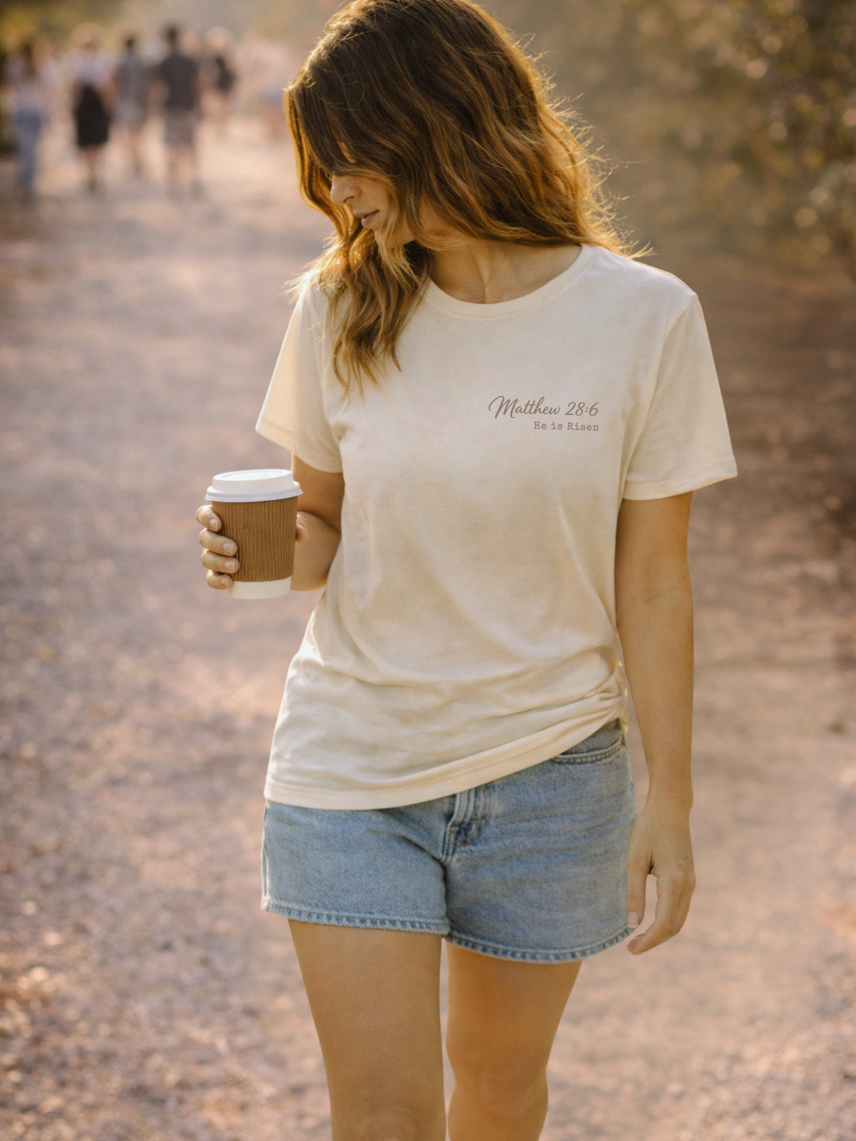 Woman wearing a natural t-shirt with text and denim shorts, holding a coffee cup on a path.