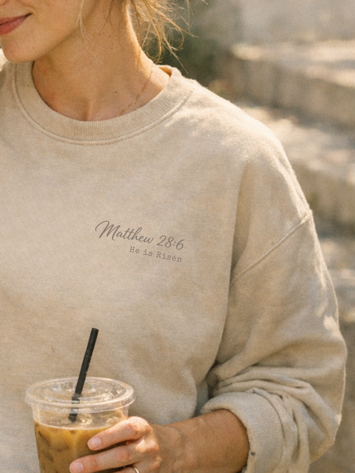 Woman wearing a beige sweatshirt with text, holding an iced coffee. Christian Matthew 28:6