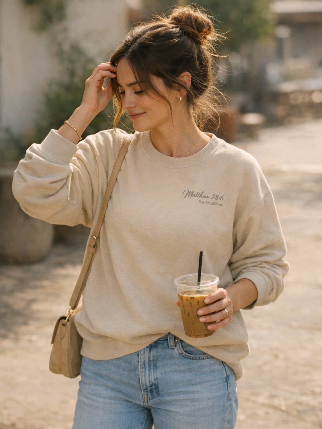 Woman in a beige sweatshirt and blue jeans holding an iced coffee, standing outdoors. Matthew 28:6