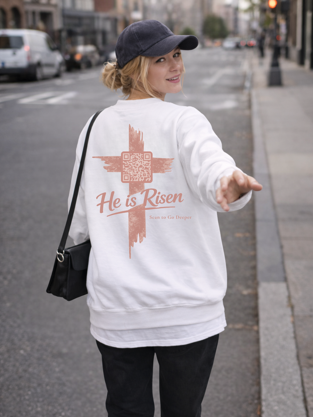 Woman wearing a white sweatshirt with 'He is Risen' text on a city street.