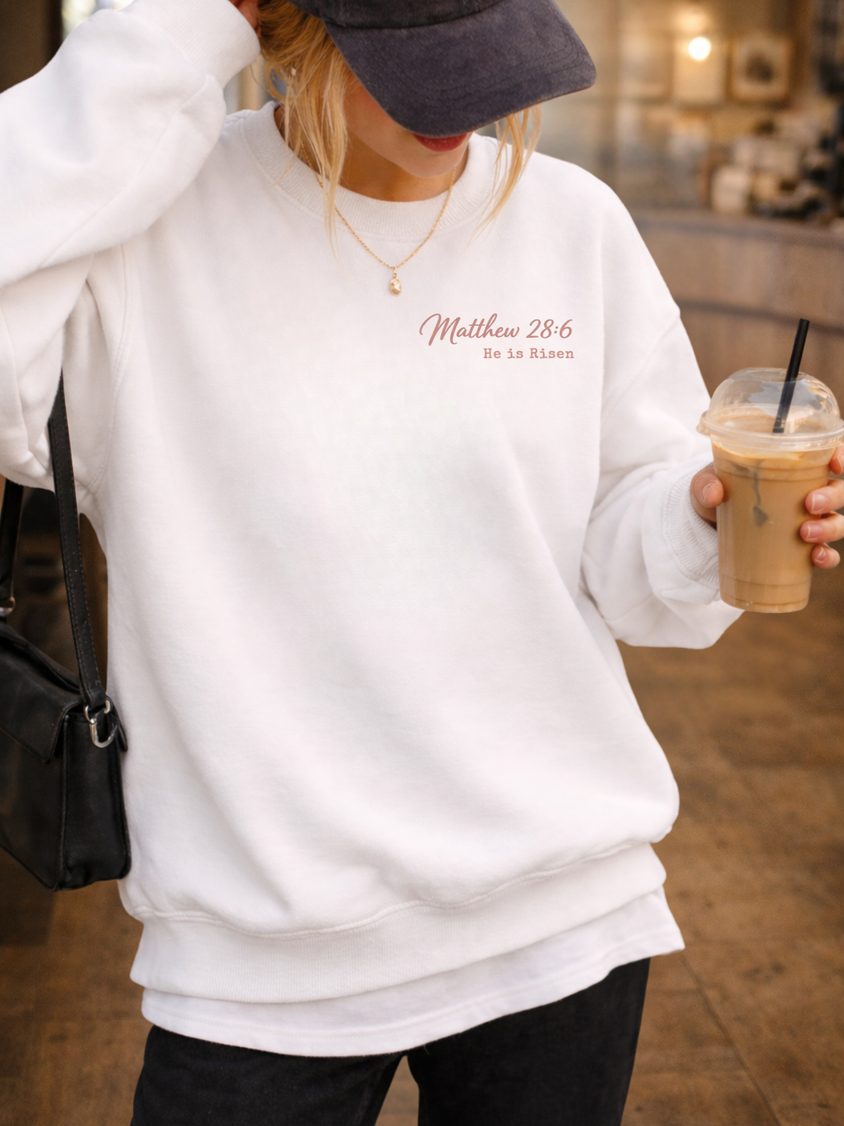 Person wearing a white sweatshirt with text, holding a coffee cup indoors. Matthew 28:6
