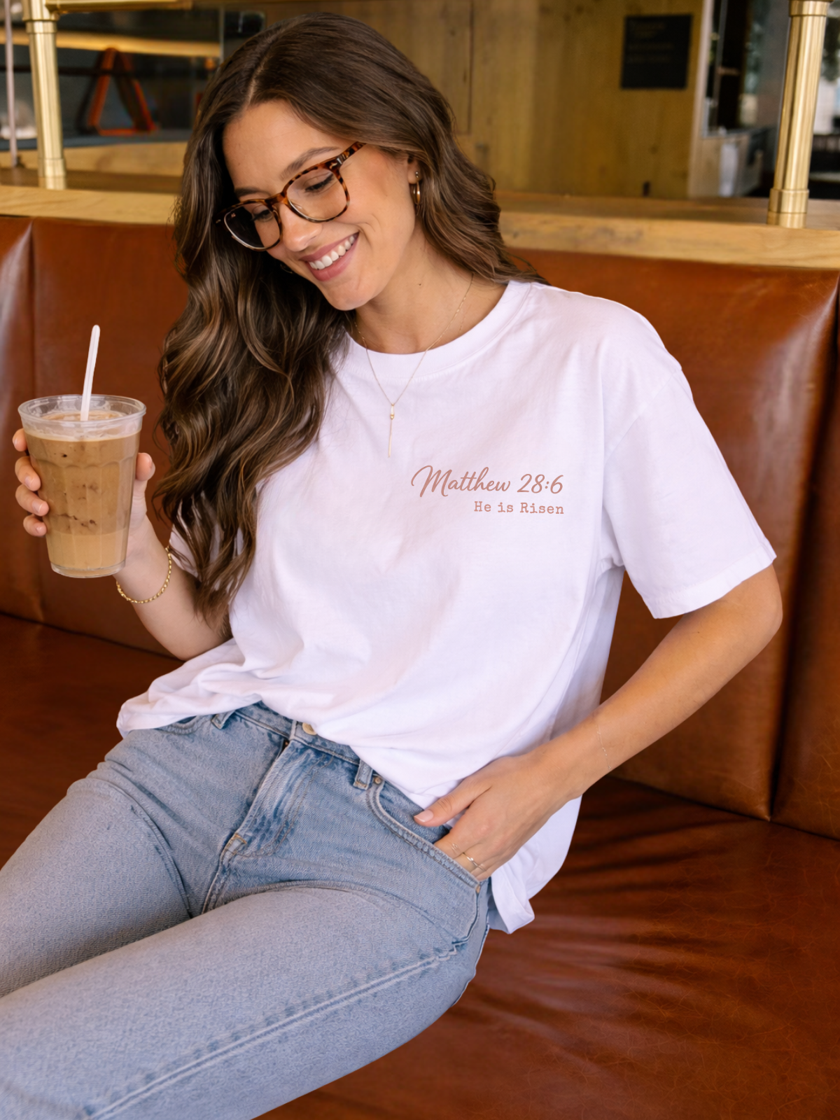 Woman holding a drink and wearing a white t-shirt with text, sitting on a brown leather couch.